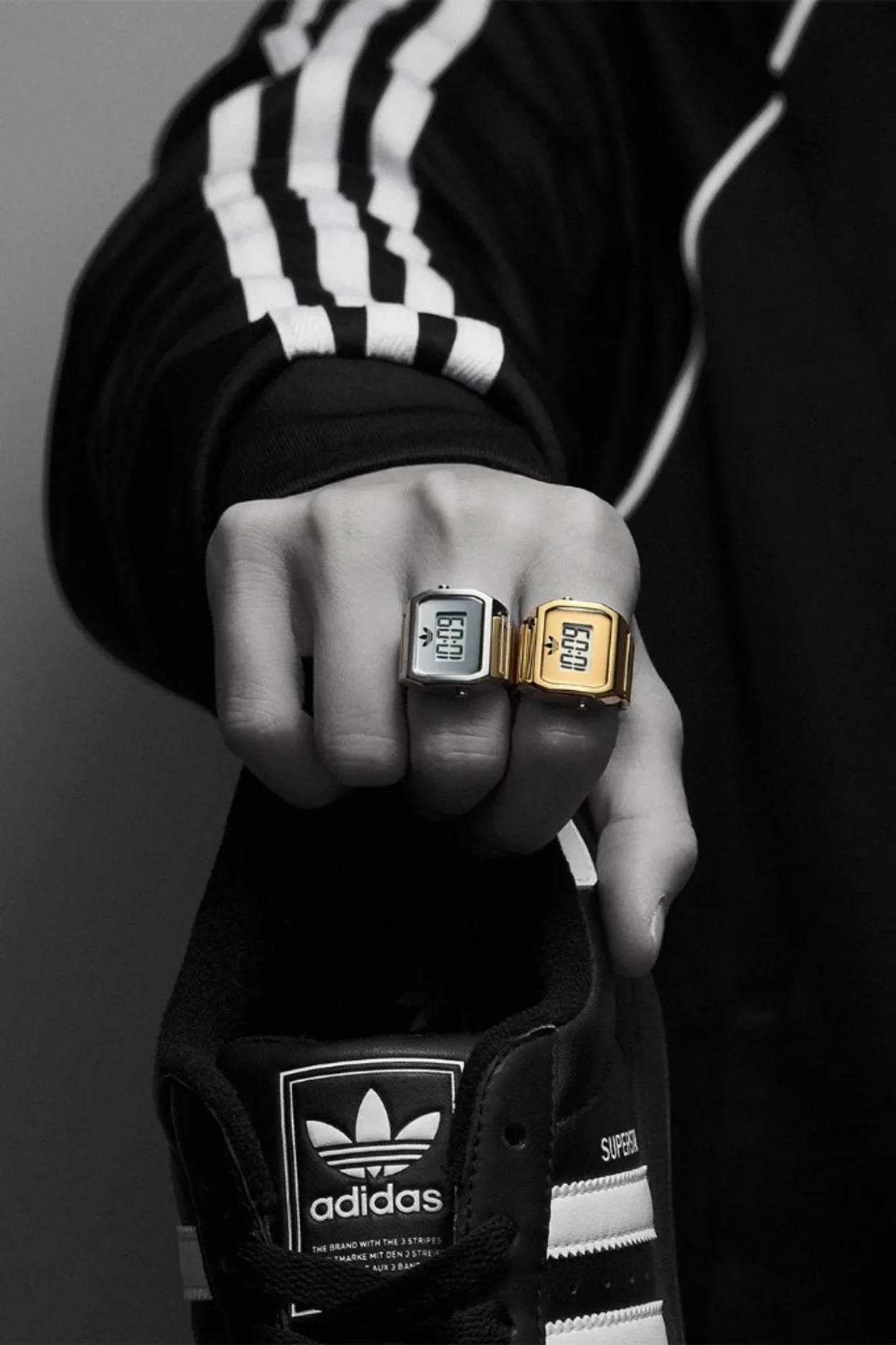 adidas Digital Two ring watches in silver and gold on a fist with a black adidas Superstar sneaker and track jacket.
