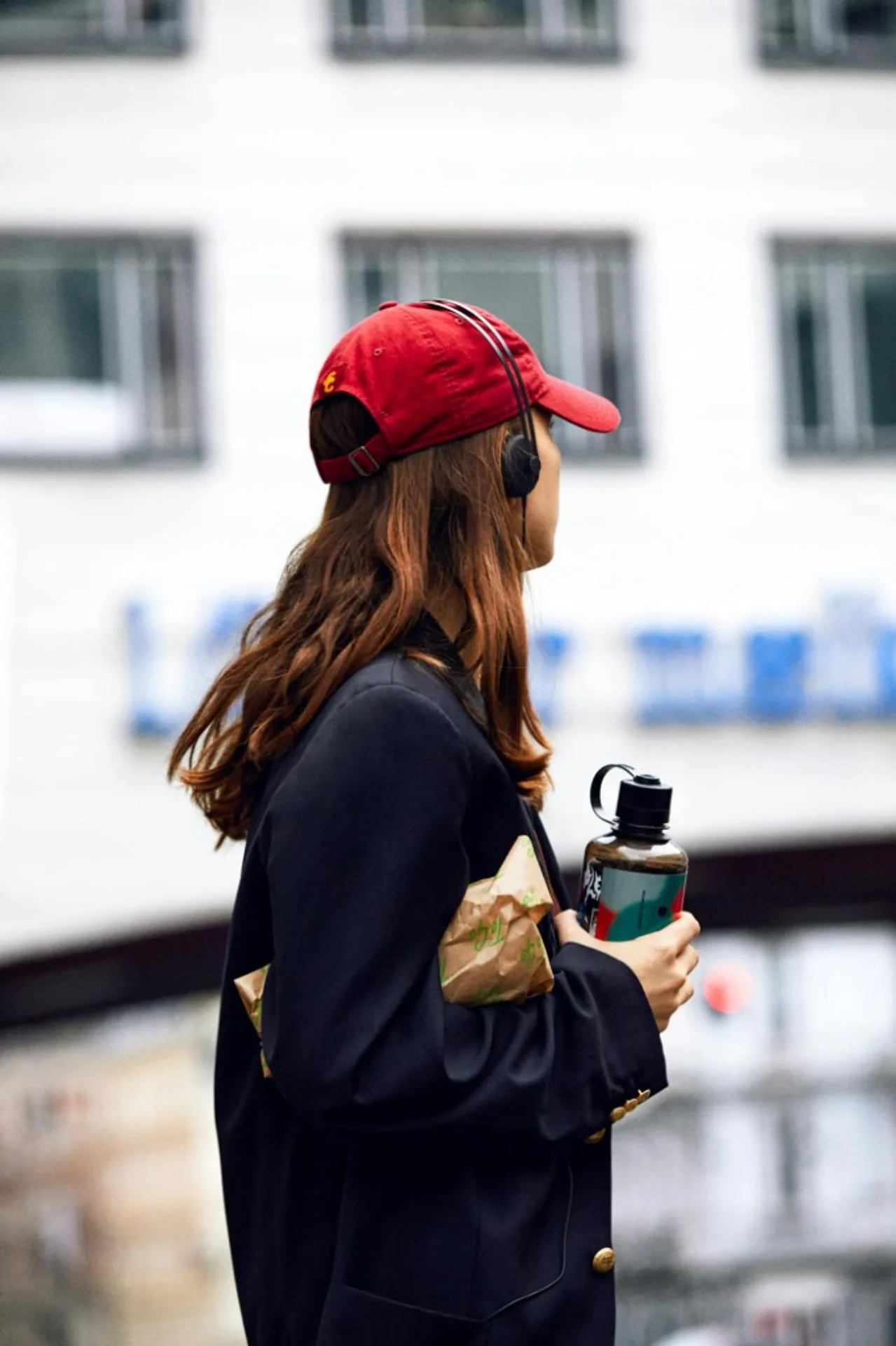 AIAIAI Tracks on-ear headphones over a red cap, side profile in navy blazer with gold buttons, urban building backdrop bokeh.