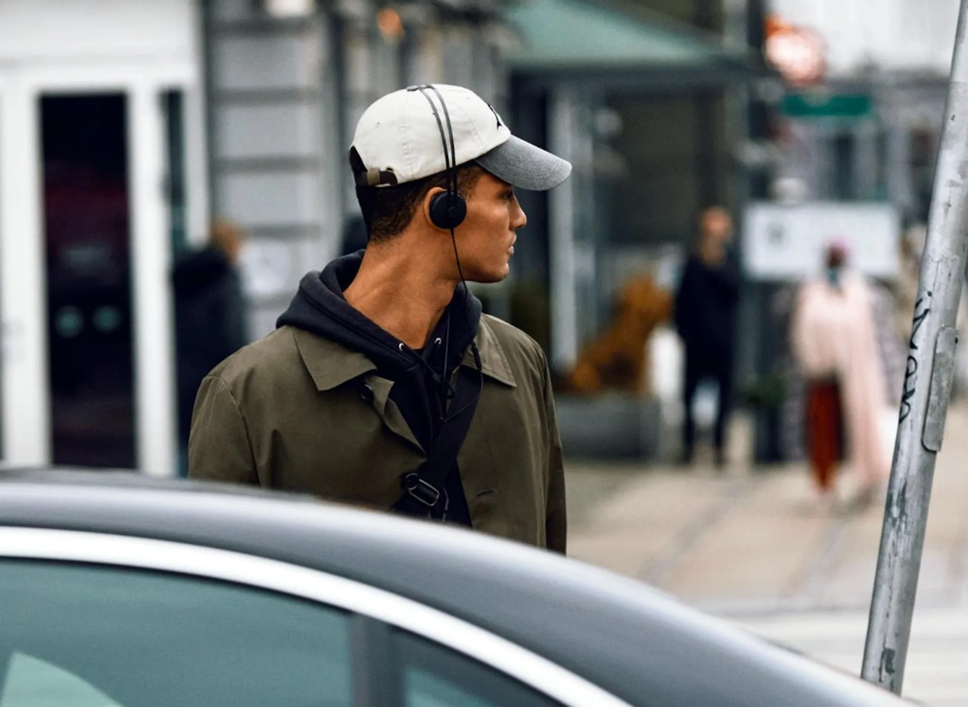 AIAIAI Tracks black on-ear headphones worn over a cap, urban street profile with shallow depth of field, olive coat and cros…
