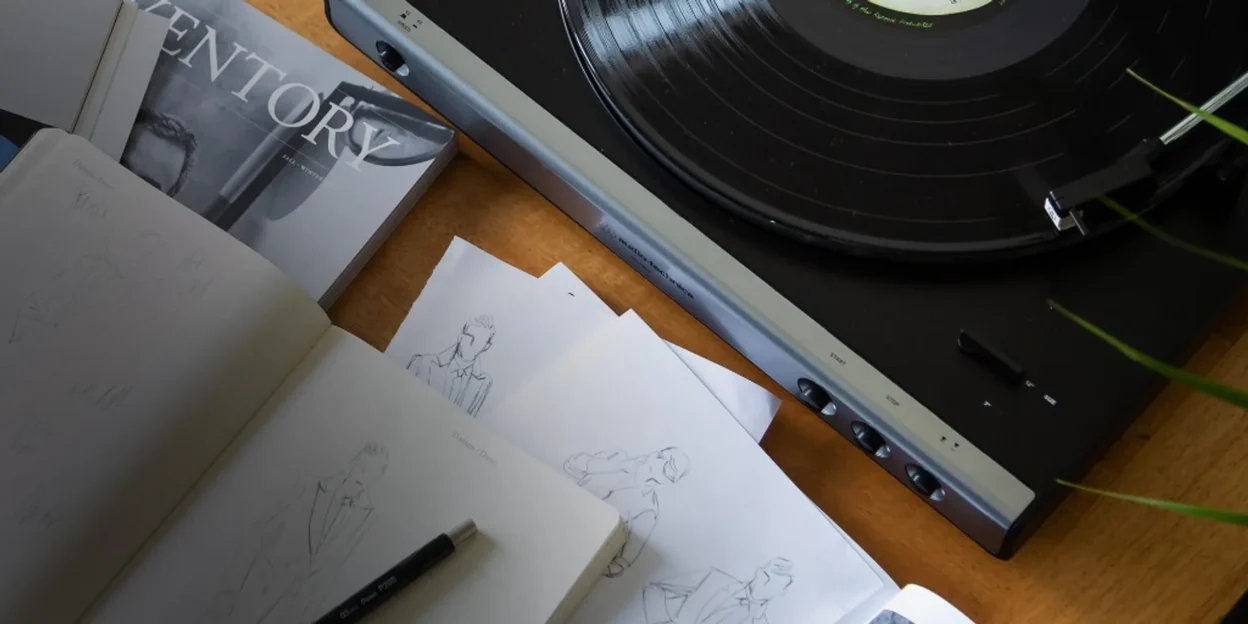 Lifestyle flat-lay: Audio-Technica turntable playing vinyl on a wooden desk with magazine, fashion sketches, and mechanical…