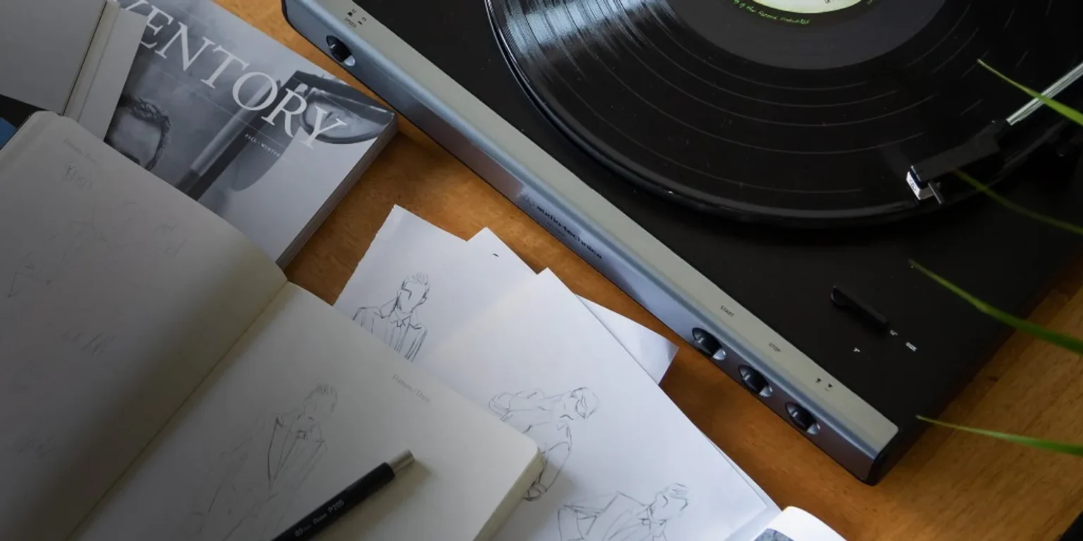 Lifestyle flat-lay: Audio-Technica turntable playing vinyl on a wooden desk with magazine, fashion sketches, and mechanical…