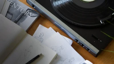 Lifestyle flat-lay: Audio-Technica turntable playing vinyl on a wooden desk with magazine, fashion sketches, and mechanical…