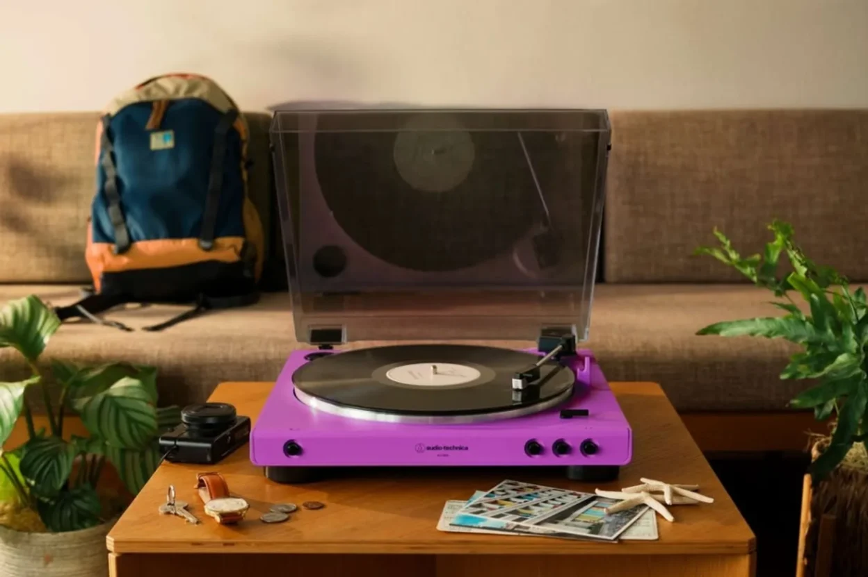 Purple Audio-Technica AT-LP60XBT turntable playing a record on a wooden table with dust cover open, lifestyle scene with sof…
