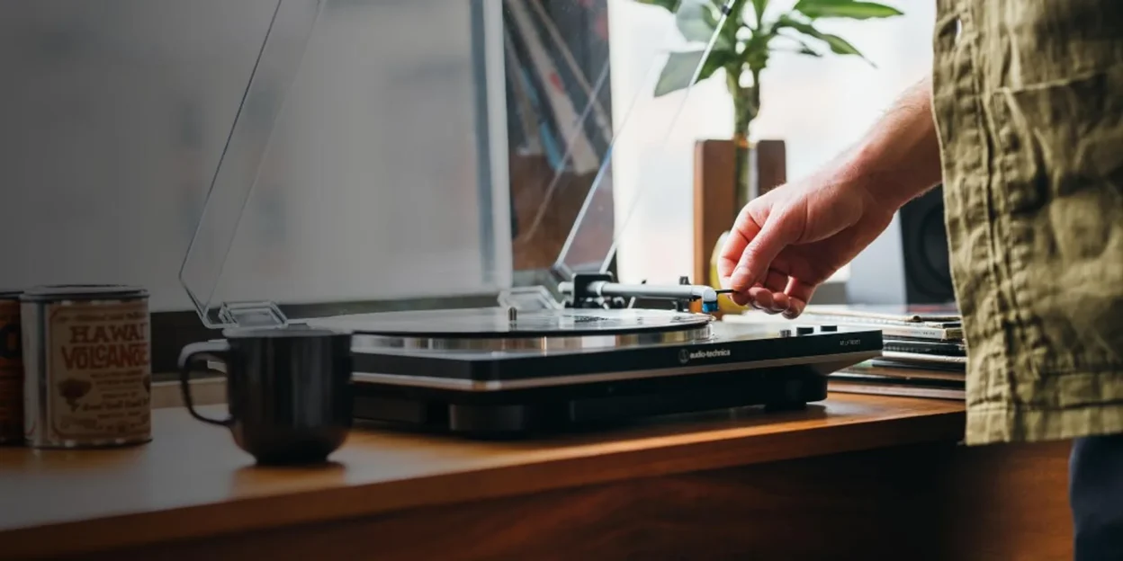 Lifestyle shot: hand lowering the tonearm on a black Audio-Technica turntable on a light wood desk with vinyl, mug and natur…