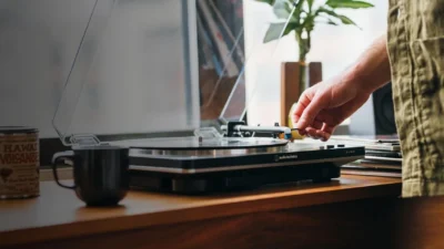 Lifestyle shot: hand lowering the tonearm on a black Audio-Technica turntable on a light wood desk with vinyl, mug and natur…