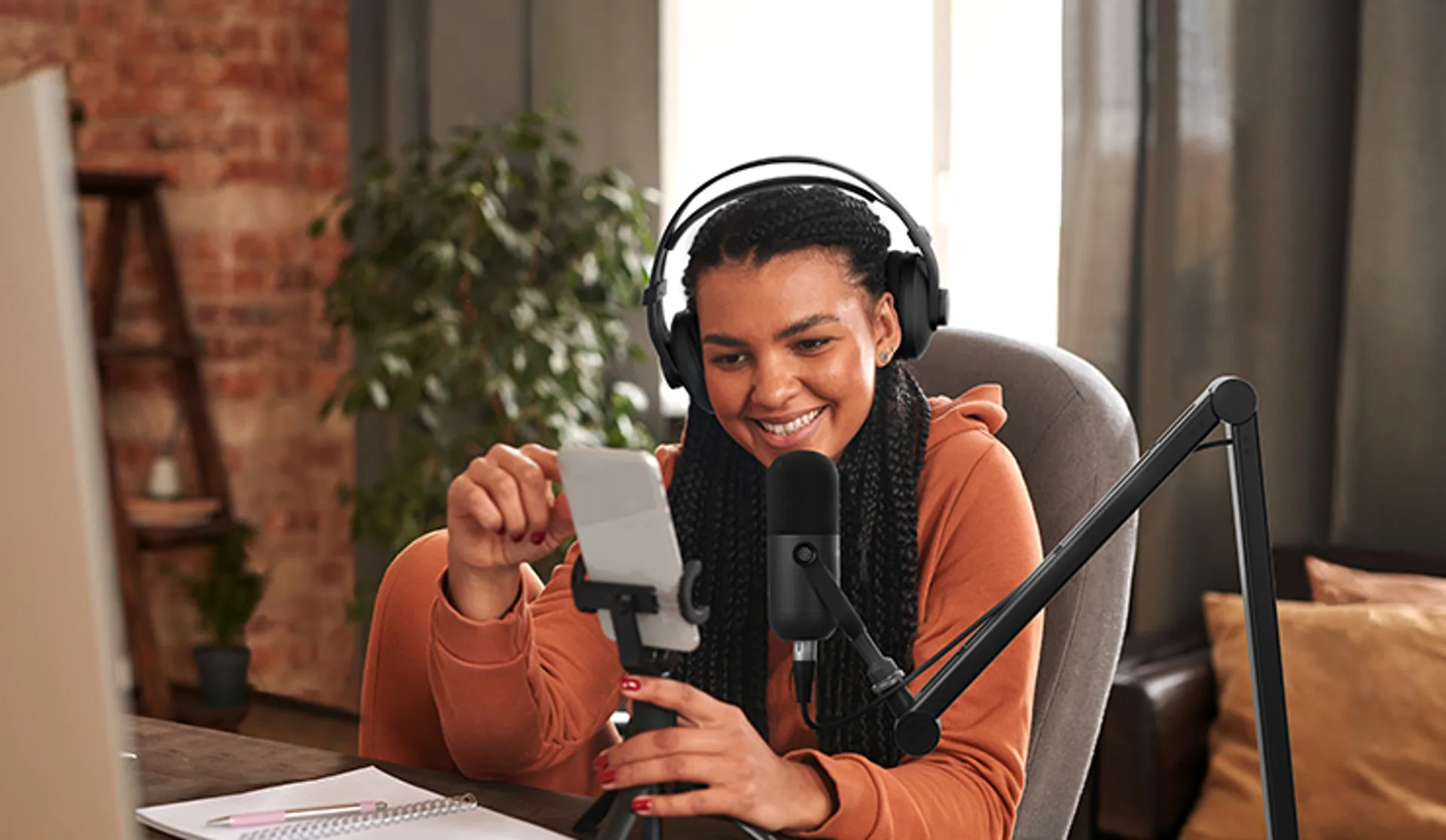 Woman at a home desk with BOYA mic on a boom arm, smartphone on a small tripod, and over-ear headphones.