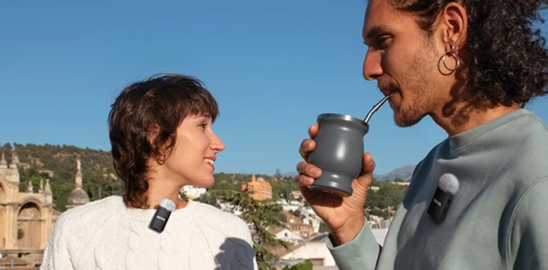 Two people outdoors with BOYA clip-on wireless microphones and windscreens, sunny city background.