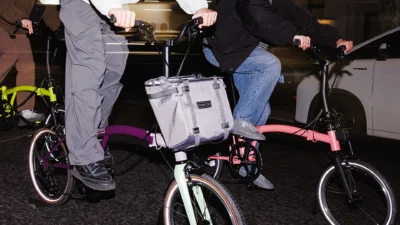 Night urban scene: riders on Brompton P Line Asia-Exclusive folding bikes with two-tone frames, tan-wall tires and front bag…