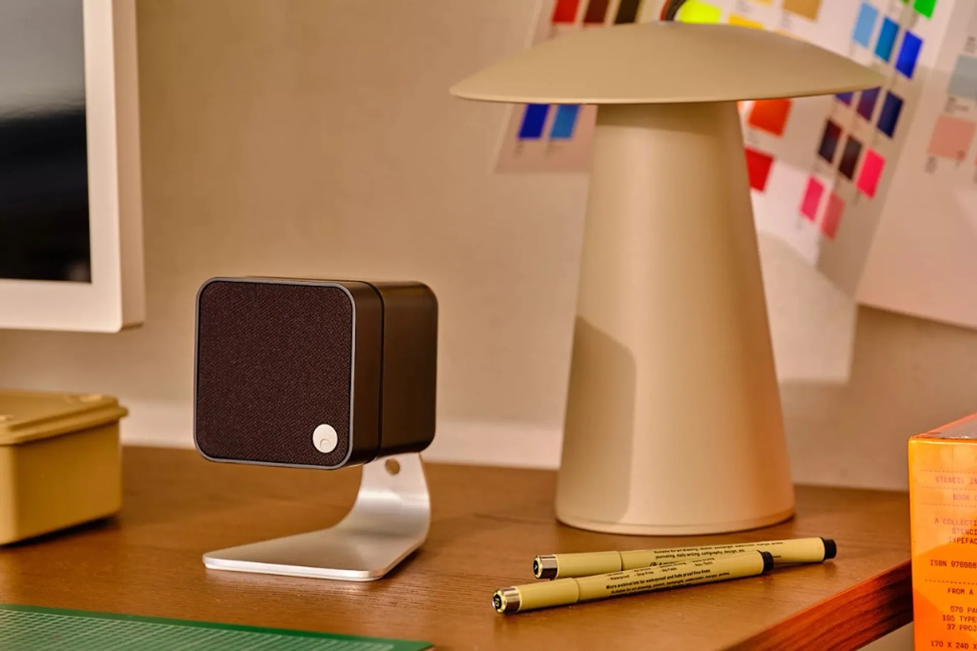 Black Cambridge Audio Minx compact speaker on a silver C-shaped desk stand on a wooden desk with mushroom lamp, monitor and…