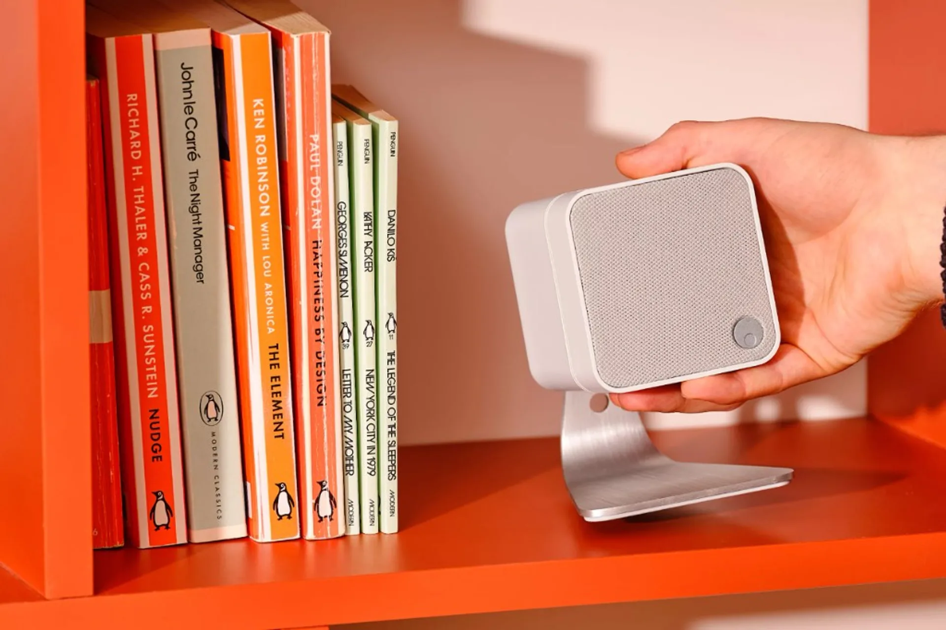Hand placing a small white Cambridge Audio Minx satellite speaker onto a brushed silver desktop stand on an orange bookshelf…