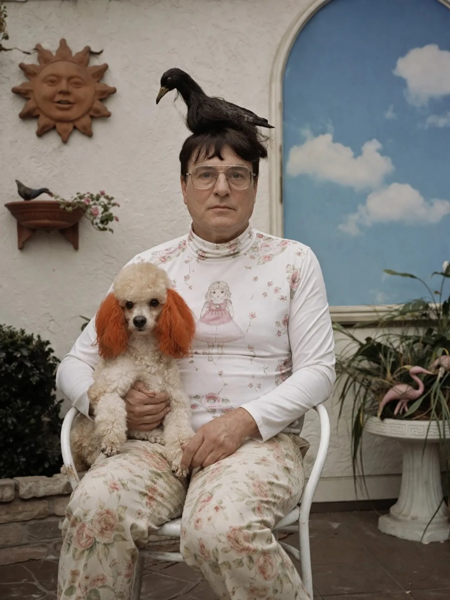 Film-style surreal portrait: man in floral outfit on patio chair with small dog on lap and black bird on head, kitschy suburban patio with flamingos.