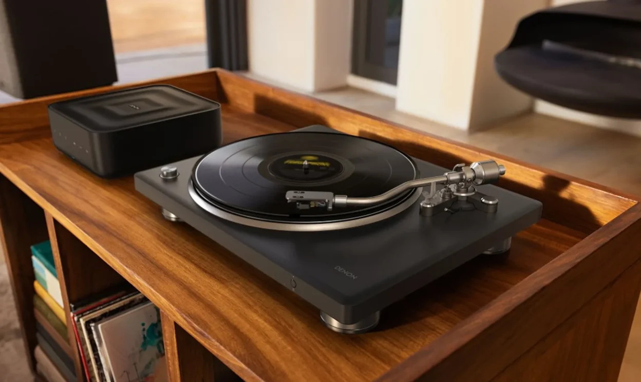 Wide lifestyle: Denon DP-500BT turntable playing vinyl on a wood cabinet beside a Denon wireless speaker; man relaxes on the…