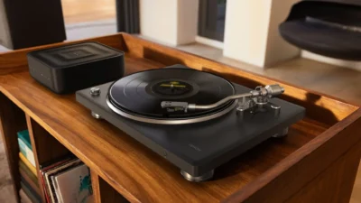 Wide lifestyle: Denon DP-500BT turntable playing vinyl on a wood cabinet beside a Denon wireless speaker; man relaxes on the…