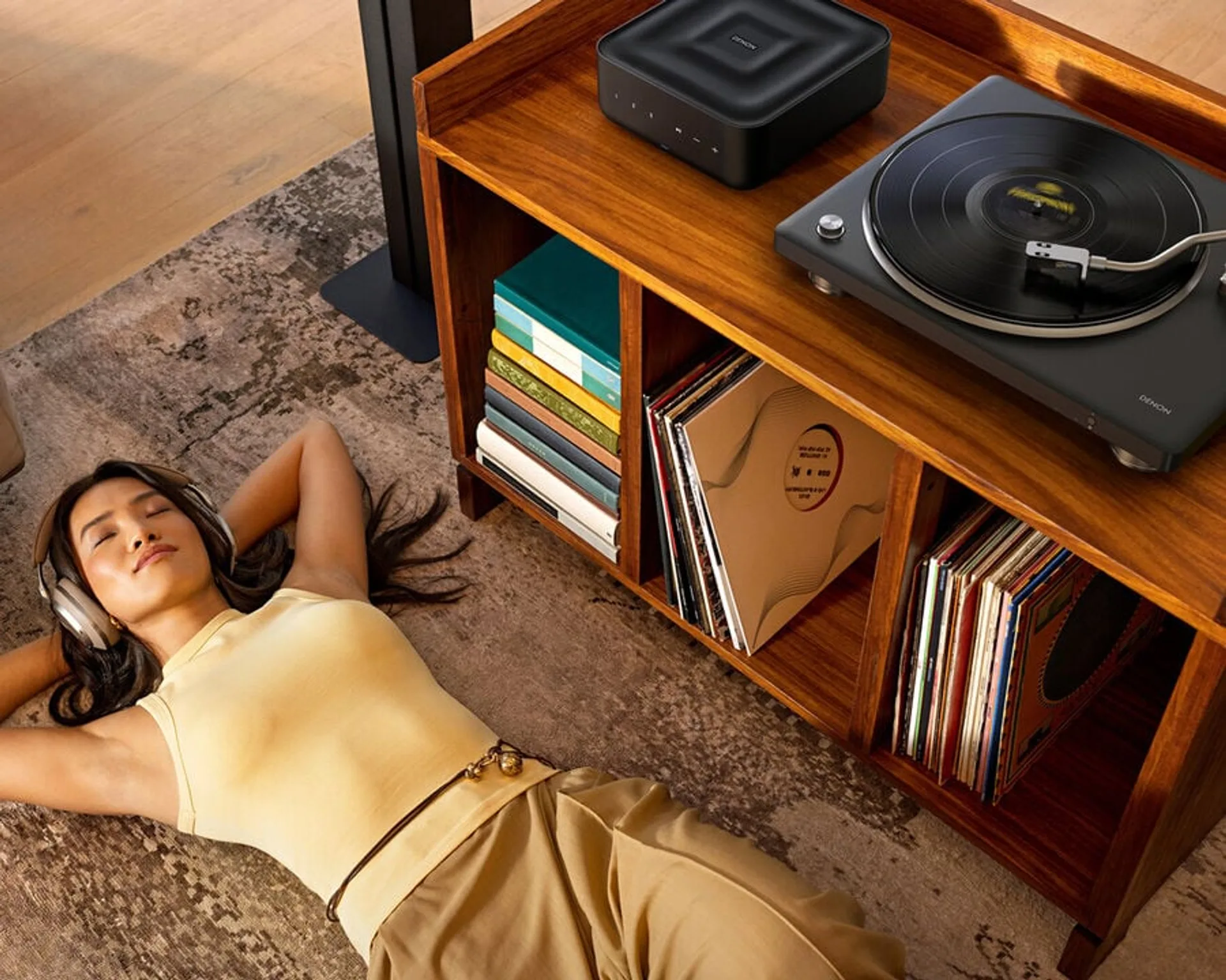 Overhead lifestyle: woman resting on a patterned rug wearing headphones, Denon DP-500BT and Denon wireless speaker on a mid-…