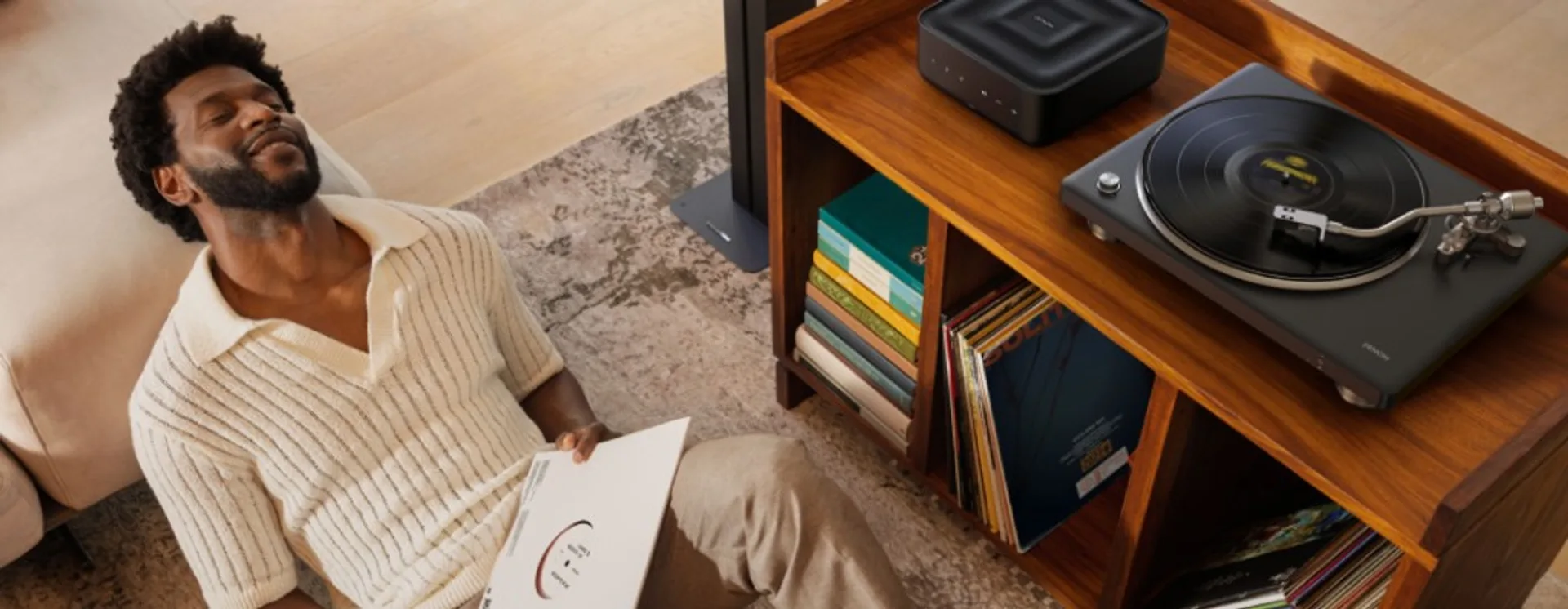 Denon DP-500BT turntable with silver tonearm playing a record next to a black Denon wireless speaker on a wooden console wit…