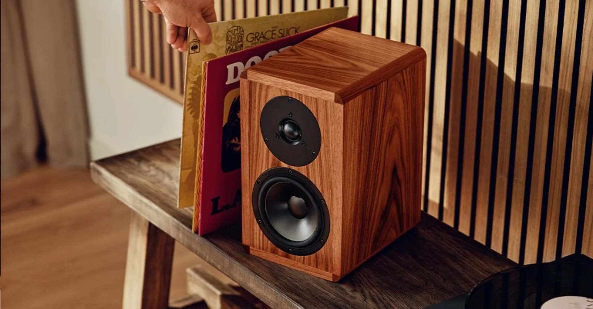 Dynaudio Heritage Special walnut speaker on wooden bench with vinyl records and hand selecting an album, vertical wood slat…