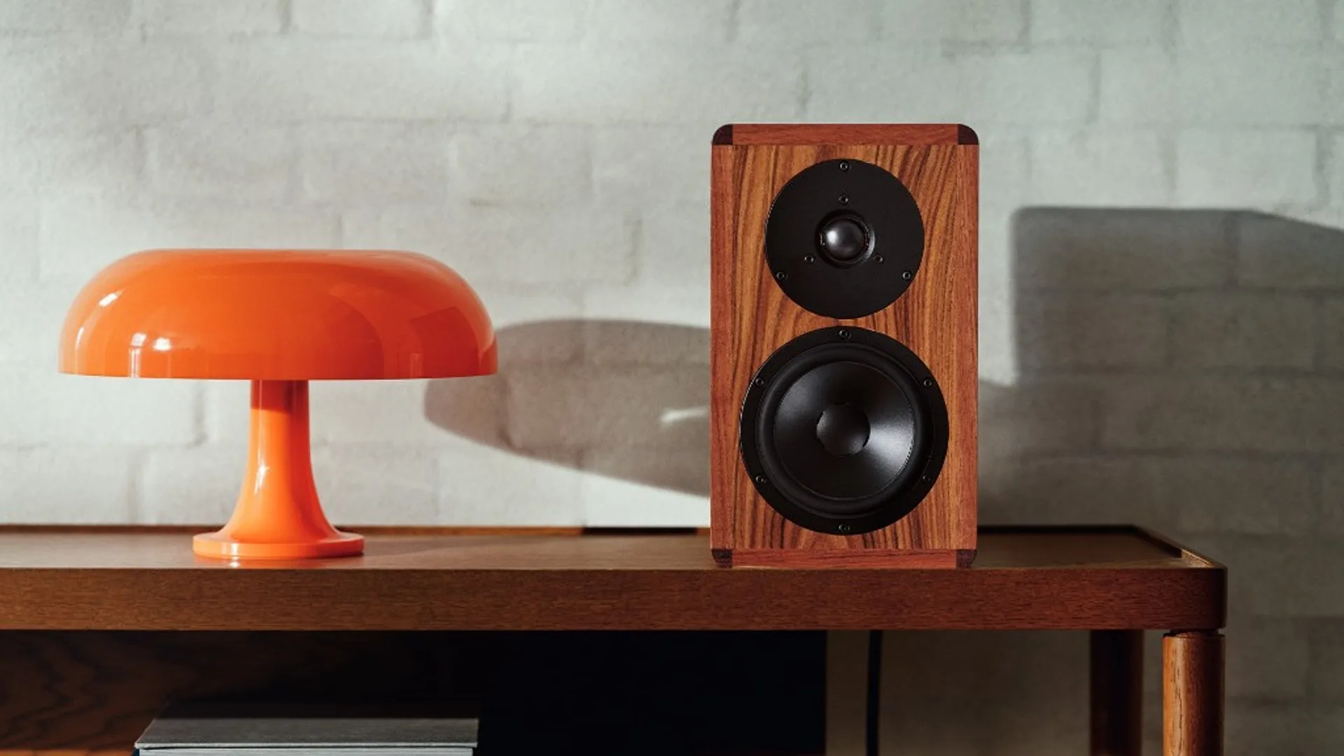 Dynaudio Heritage Special walnut bookshelf speaker on dark wood console beside an orange mushroom lamp, white brick wall, au…