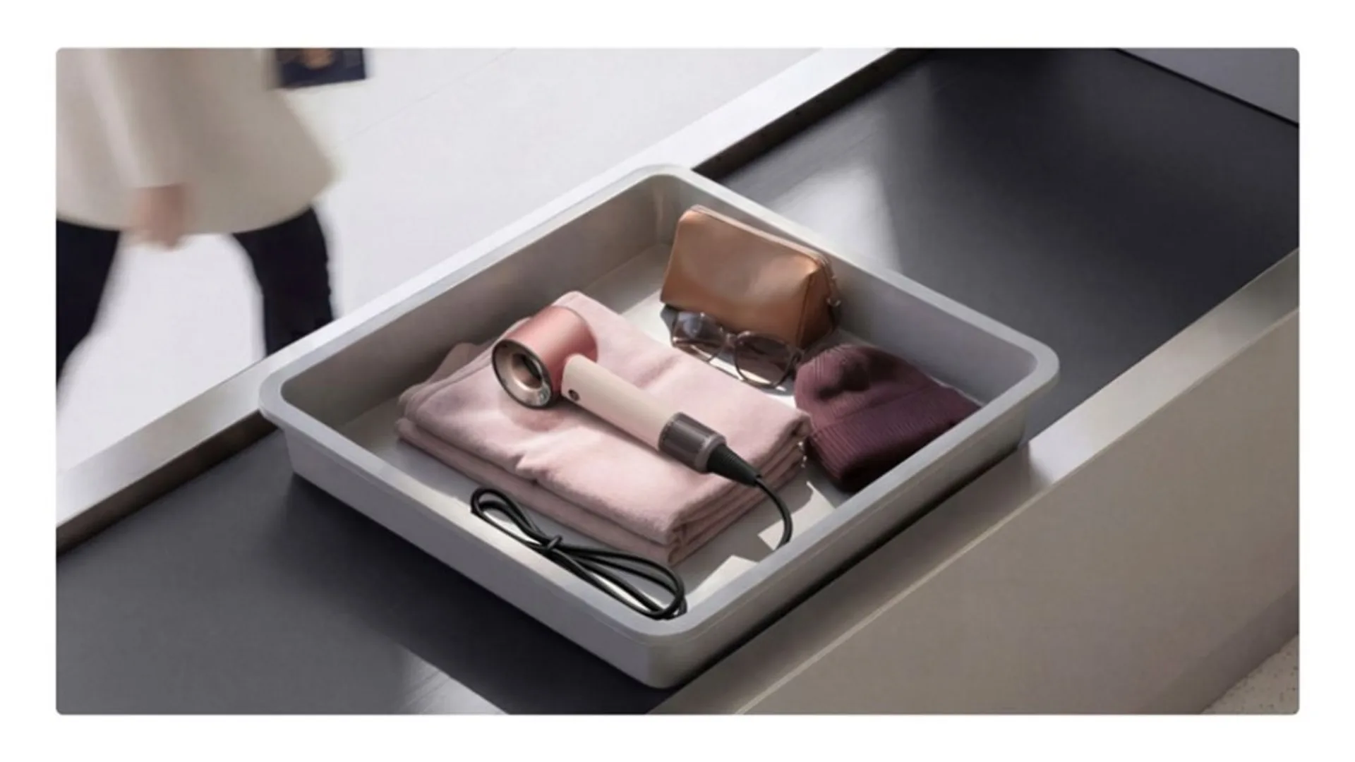 Ceramic Pink Dyson Supersonic hair dryer in a grey airport security bin with sunglasses, pouch, and folded pink clothing.