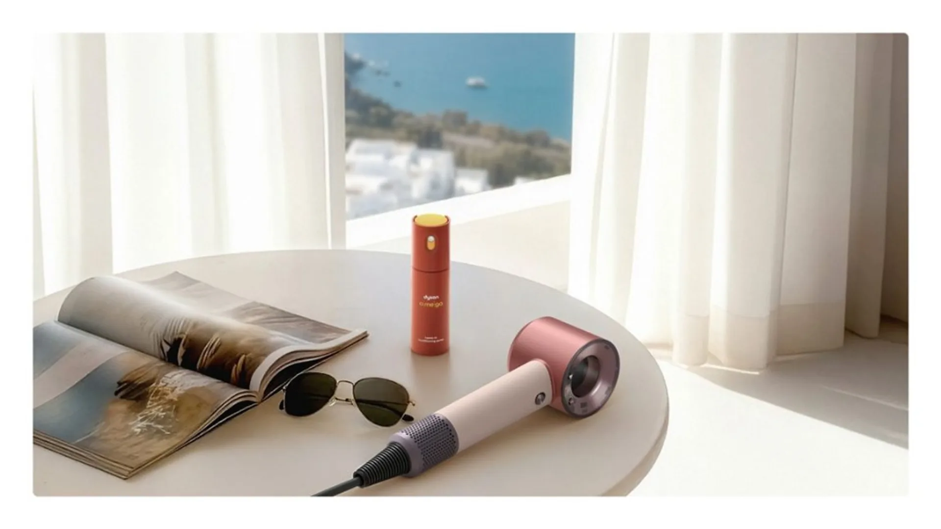 Dyson Supersonic Travel Ceramic Pink on a white table with Dyson Chime spray, sunglasses, and magazine near a bright coastal window.