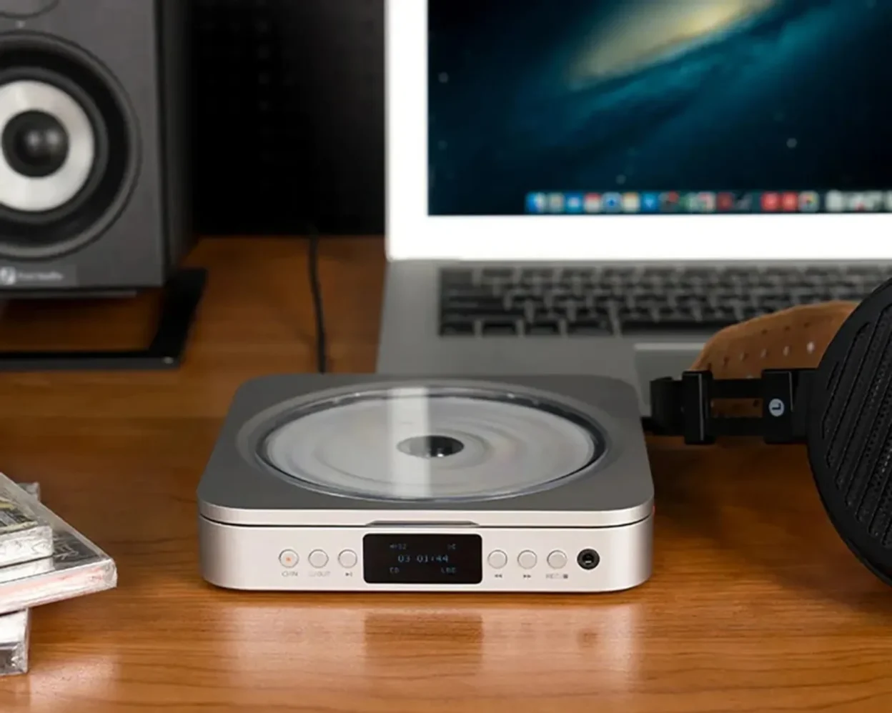 Fosi Audio Merak CD player on a wood desk beside a laptop, studio monitor speaker, stacked CDs and over-ear headphones.