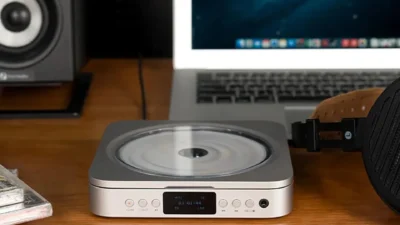 Fosi Audio Merak CD player on a wood desk beside a laptop, studio monitor speaker, stacked CDs and over-ear headphones.