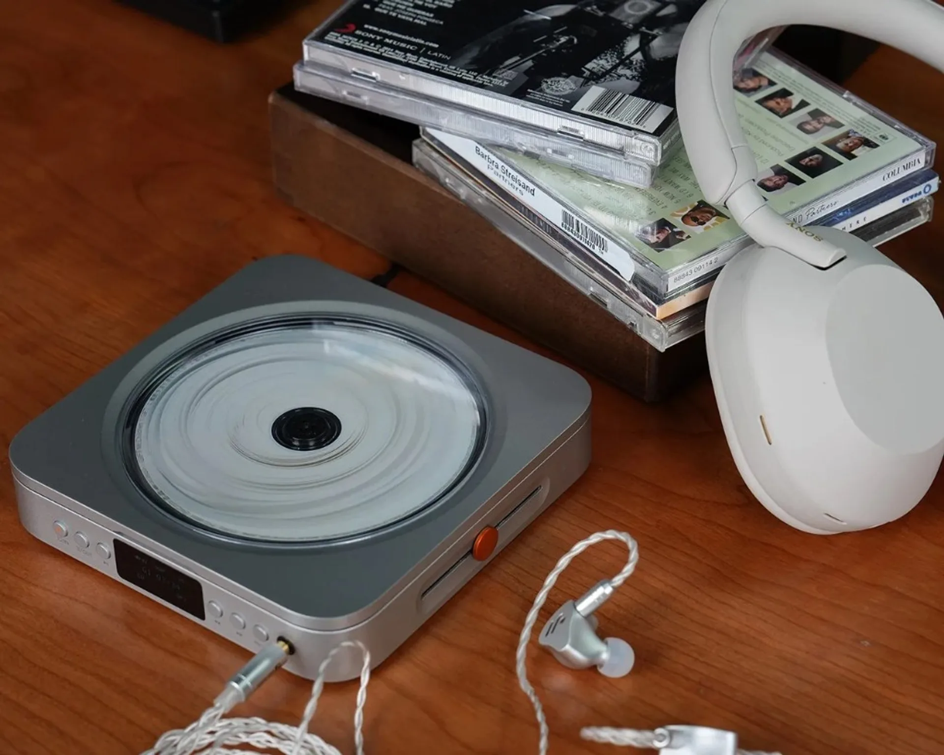 Fosi Audio Merak compact CD player on a warm wood desk with over-ear headphones, IEMs and CD jewel cases in a minimalist lis…