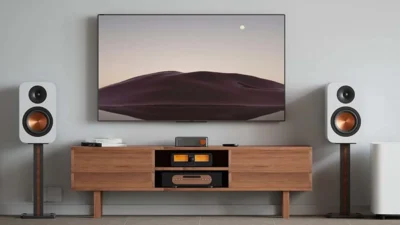 Phòng khách với TV treo tường, kệ gỗ và Fosi Audio S3 cùng loa bookshelf trắng trên chân