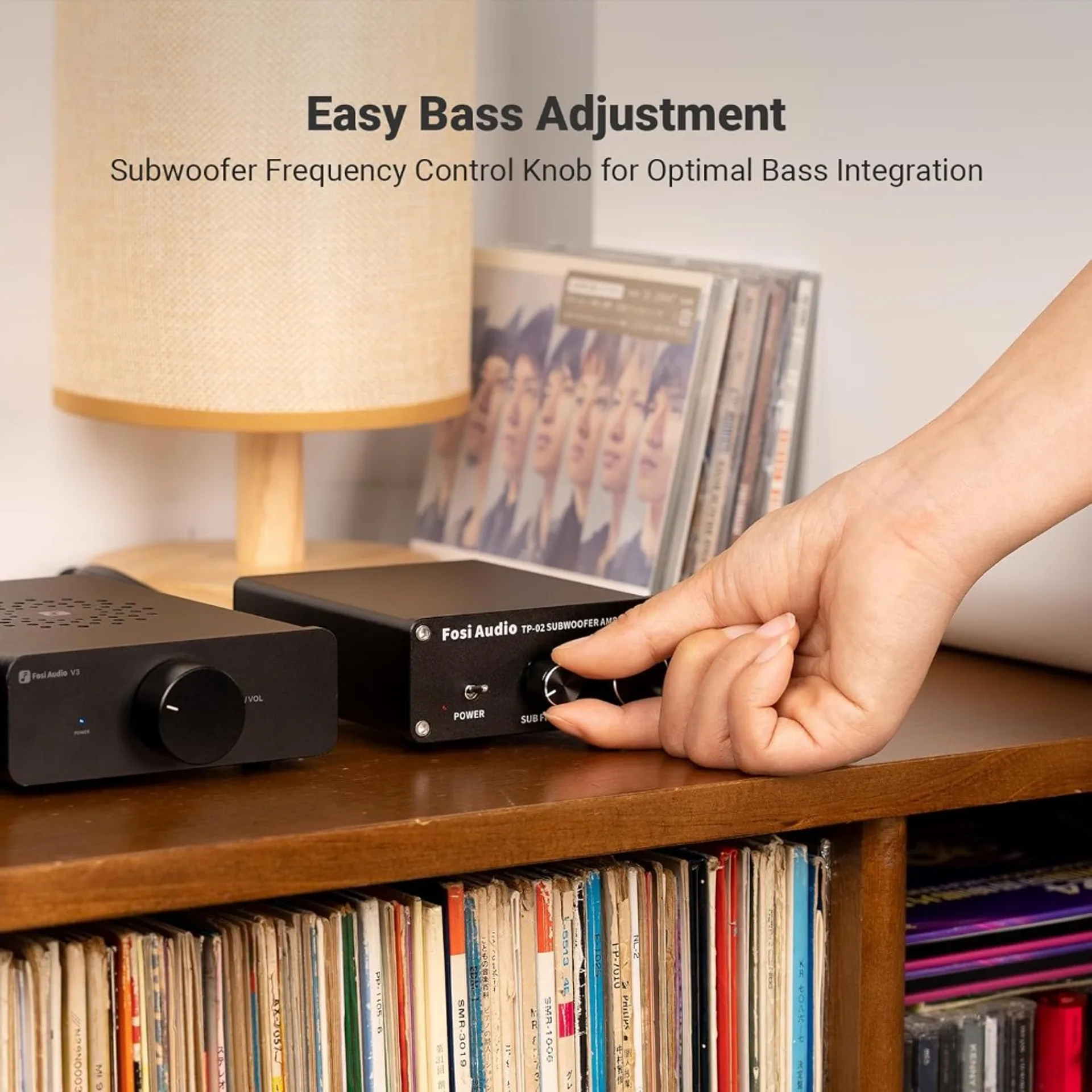 Hand adjusting sub frequency on Fosi Audio TP-02 subwoofer amp beside Fosi Audio V3 on wooden shelf with vinyl, CDs and warm…