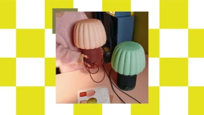 Lifestyle scene with peach and mint green IKEA KULGLASS mushroom speaker lamps on a pink table, checkerboard frame, hands ho…