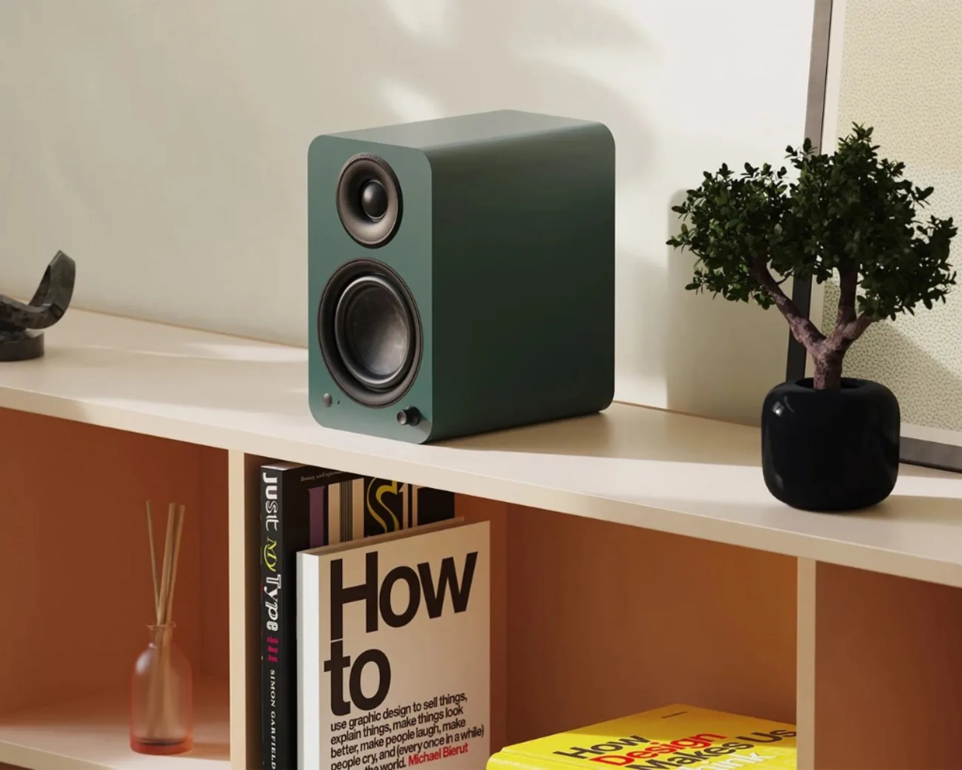 Matte forest green Kanto bookshelf speaker on a light shelf with books and small plant, soft natural light.