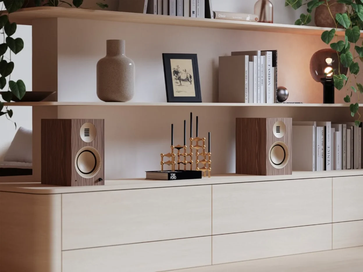 Pair of Kanto TUK Grand speakers in walnut and gold on a light wood sideboard with shelves, books, and plants.