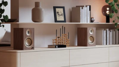 Pair of Kanto TUK Grand speakers in walnut and gold on a light wood sideboard with shelves, books, and plants.