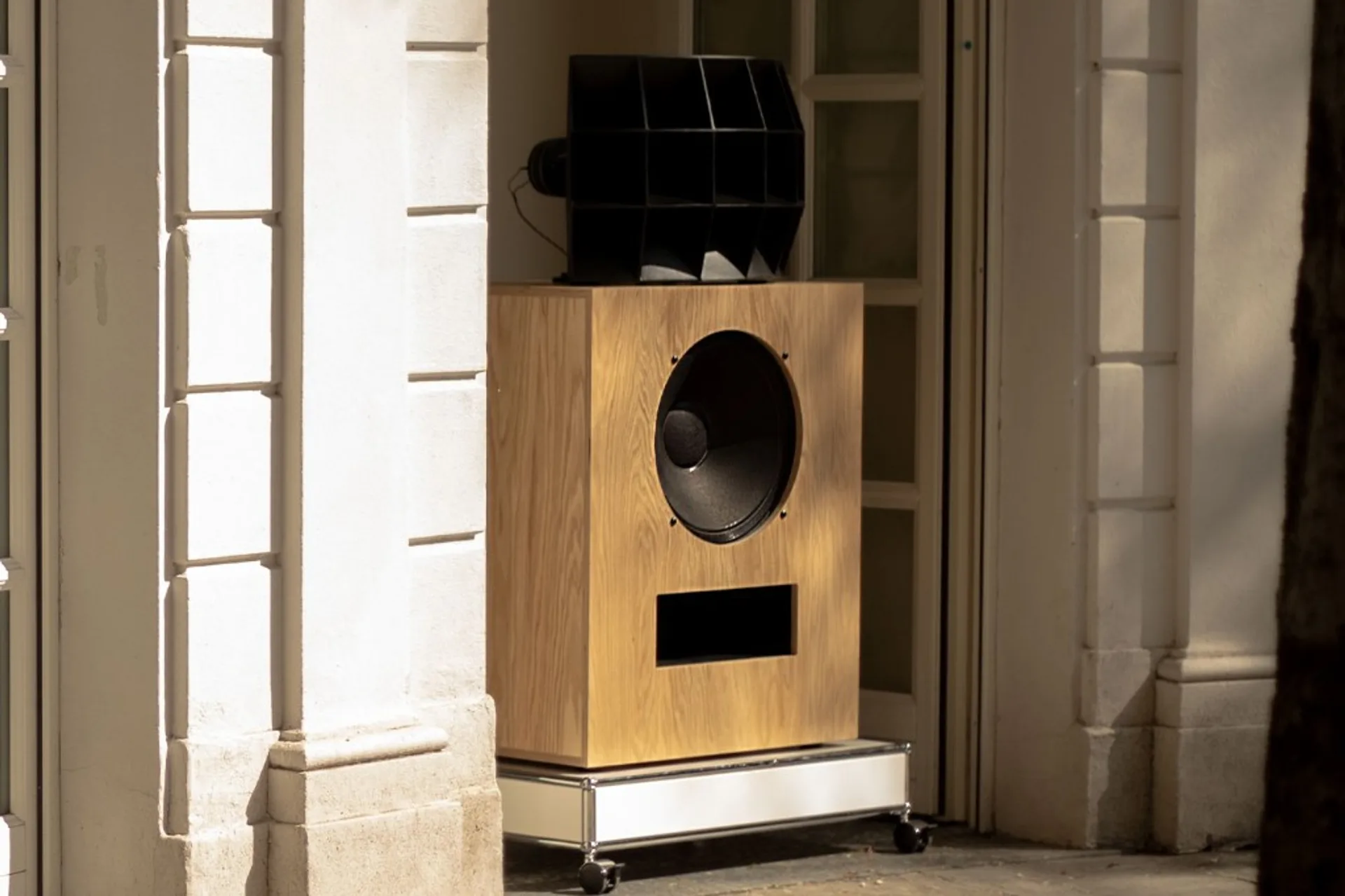 Klipsch Ojas kO-R2 loudspeaker with light wood cabinet and black multicell horn on a wheeled base in a sunlit classical door…