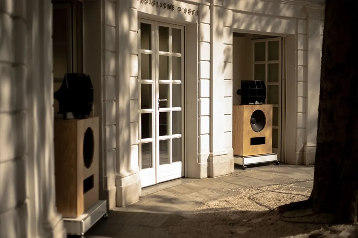 Pair of Klipsch x Ojas kO-R2 horn speakers in light wood cabinets on castored bases outside a neoclassical building inscribe…