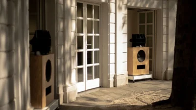 Pair of Klipsch x Ojas kO-R2 horn speakers in light wood cabinets on castored bases outside a neoclassical building inscribe…