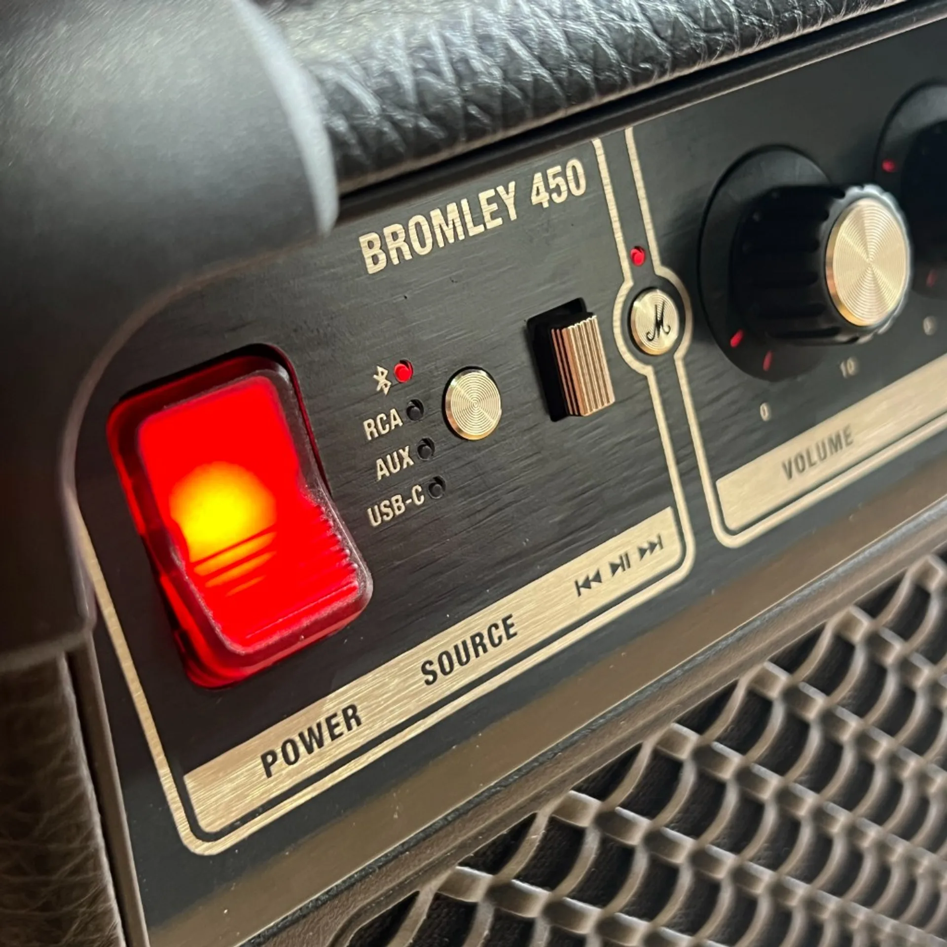 Close-up of Marshall Bromley 450 control panel: glowing red power switch, gold-accented Bluetooth RCA AUX USB-C source labels, and volume knob.