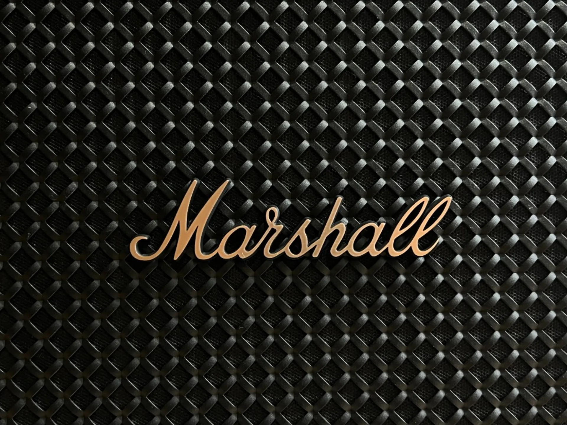 Close-up of brushed gold Marshall script logo on black diamond-weave speaker grille.