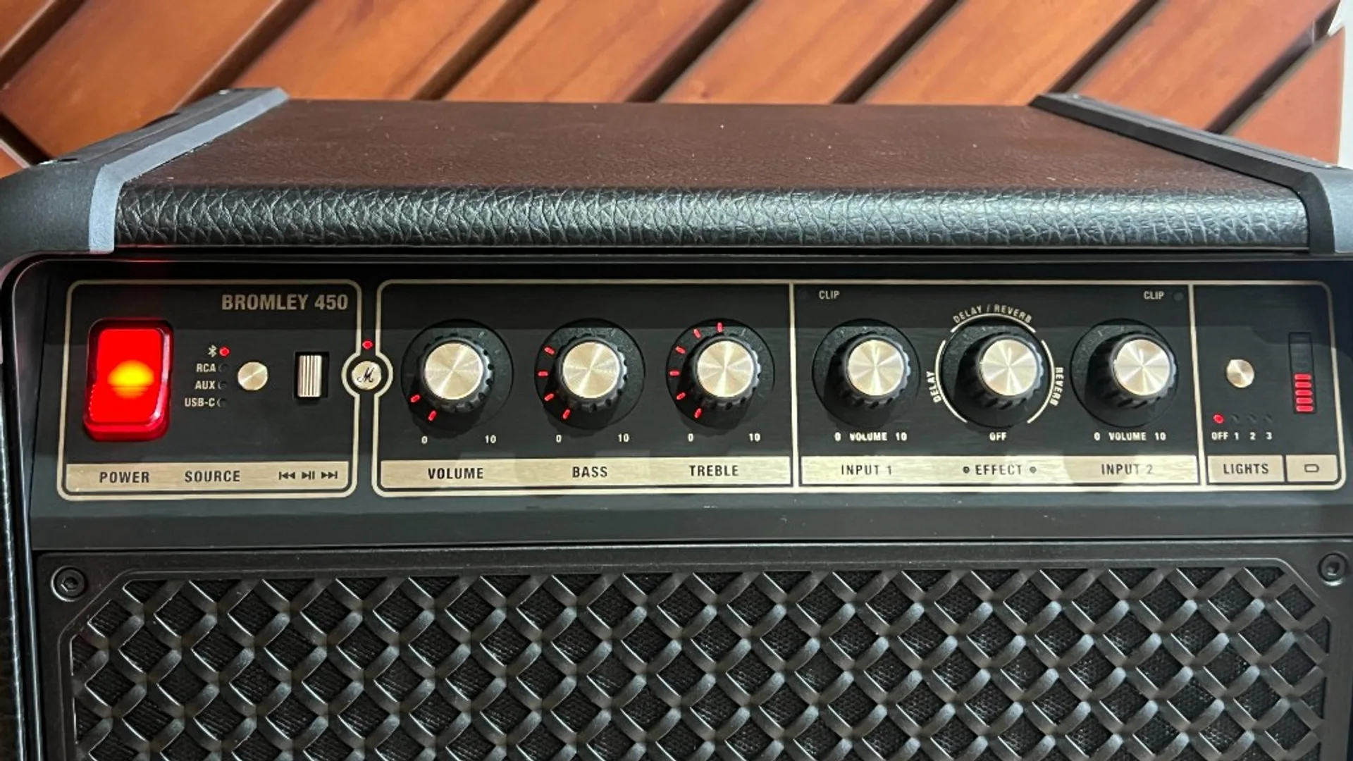 Marshall Bromley 450 top control panel: red power switch, source and playback controls, six knobs for volume bass treble inputs and delay/reverb effect, lights preset buttons, wood background.