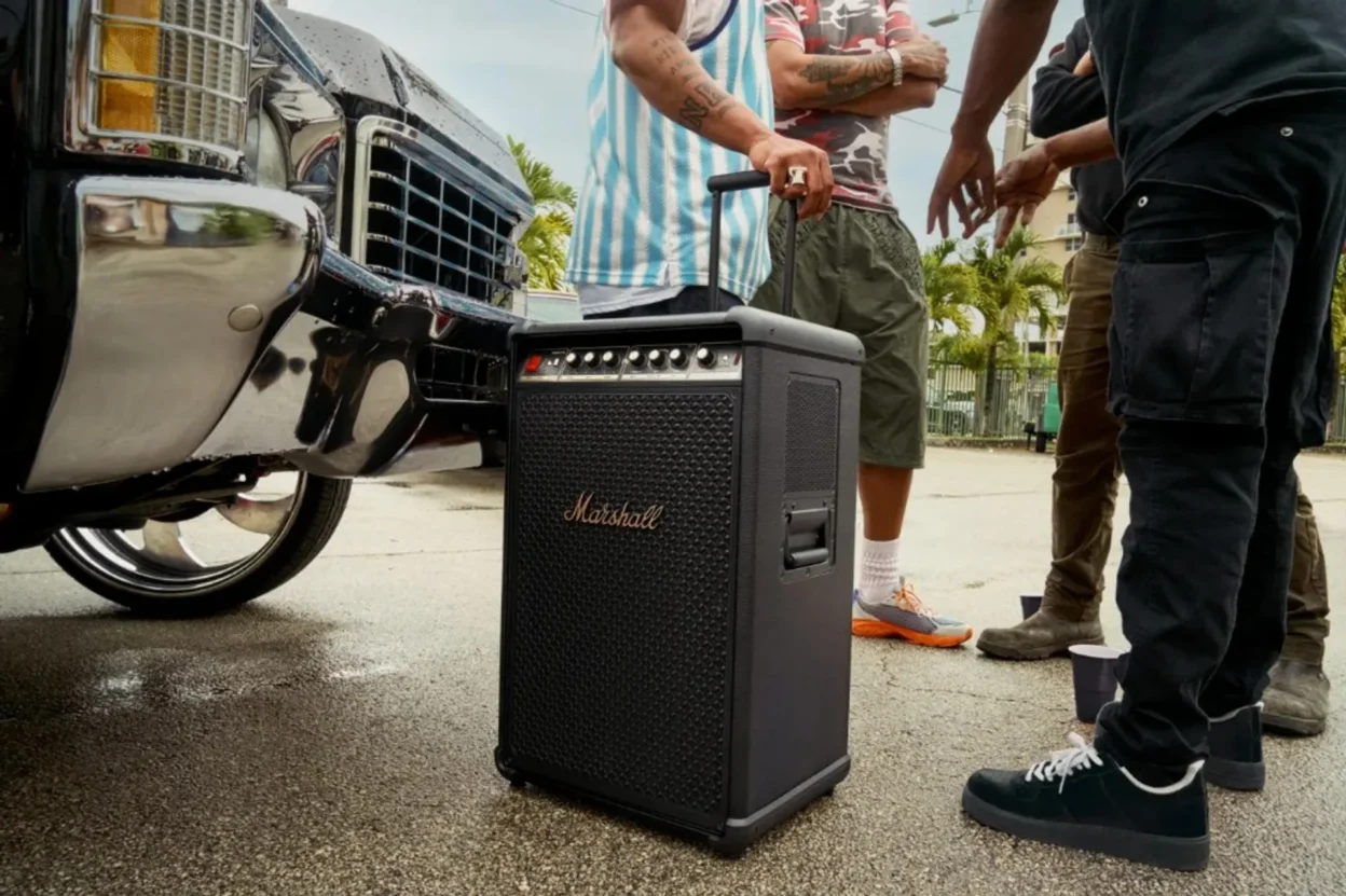 Person pulling a large black Marshall portable speaker with telescopic handle past a classic silver car, wet pavement and wa…