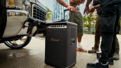 Person pulling a large black Marshall portable speaker with telescopic handle past a classic silver car, wet pavement and wa…