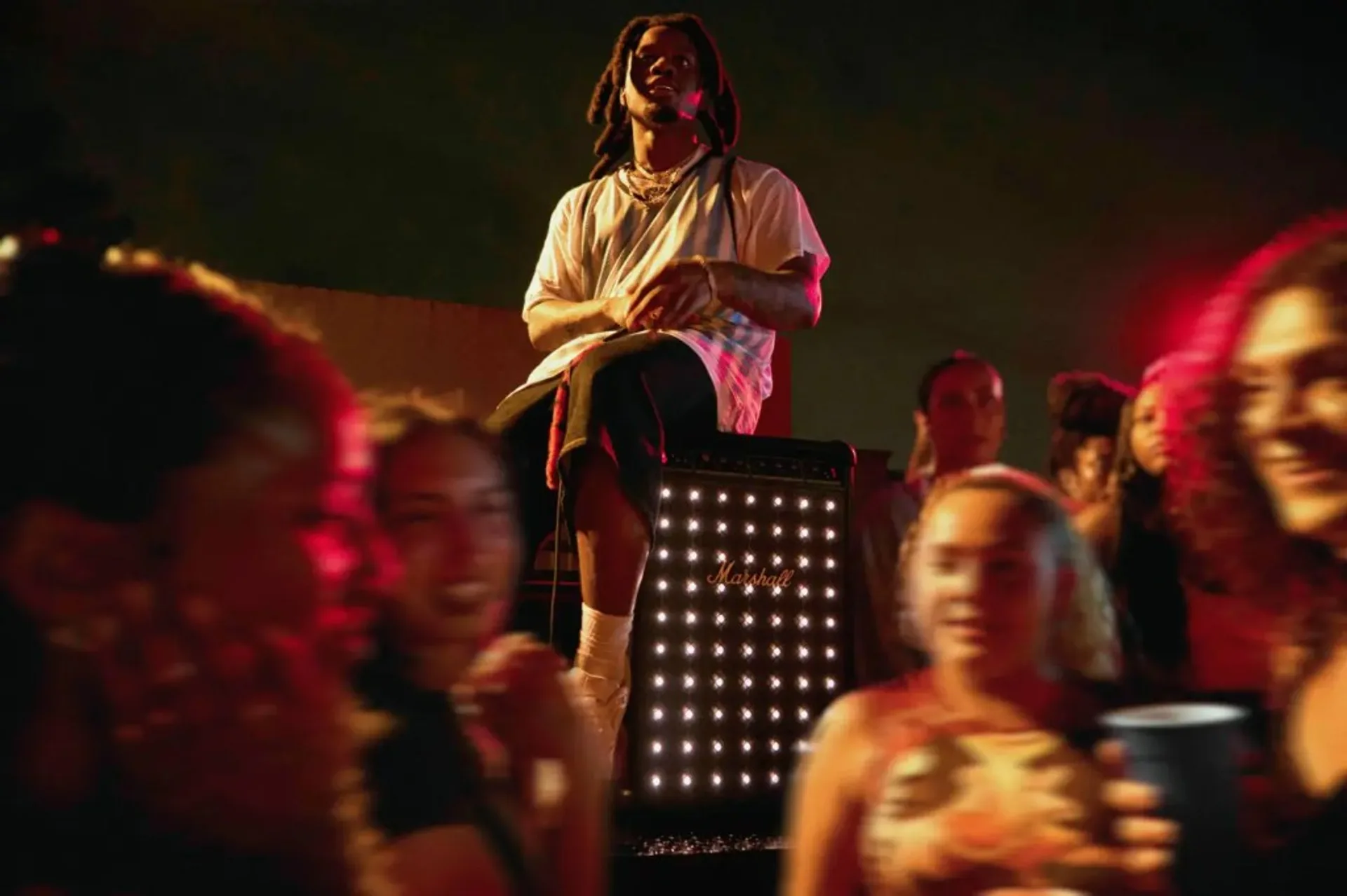 Nighttime crowd scene: person sitting on a tall Marshall speaker with illuminated dot-matrix style grille and gold logo, red…