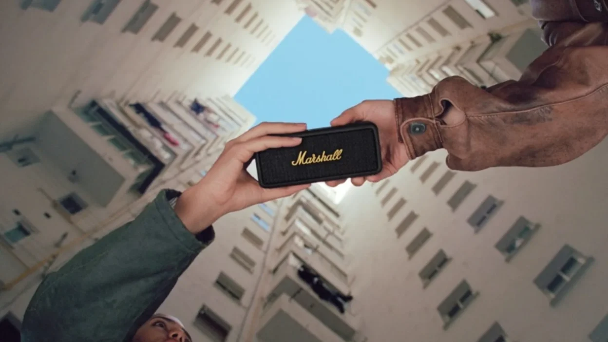 Low angle urban lifestyle shot of two people holding a black Marshall Emberton speaker against apartment buildings and blue…