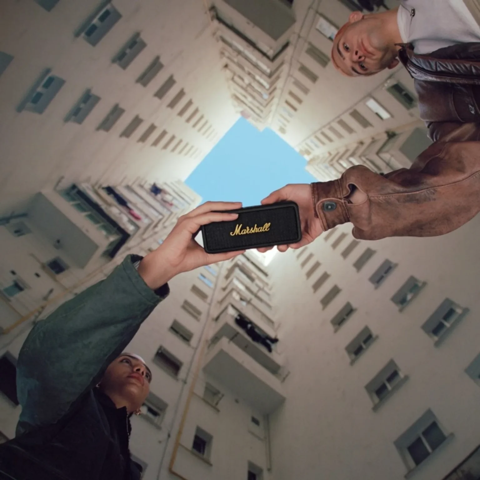 Low angle urban lifestyle shot of two people holding a black Marshall Emberton speaker against apartment buildings and blue…