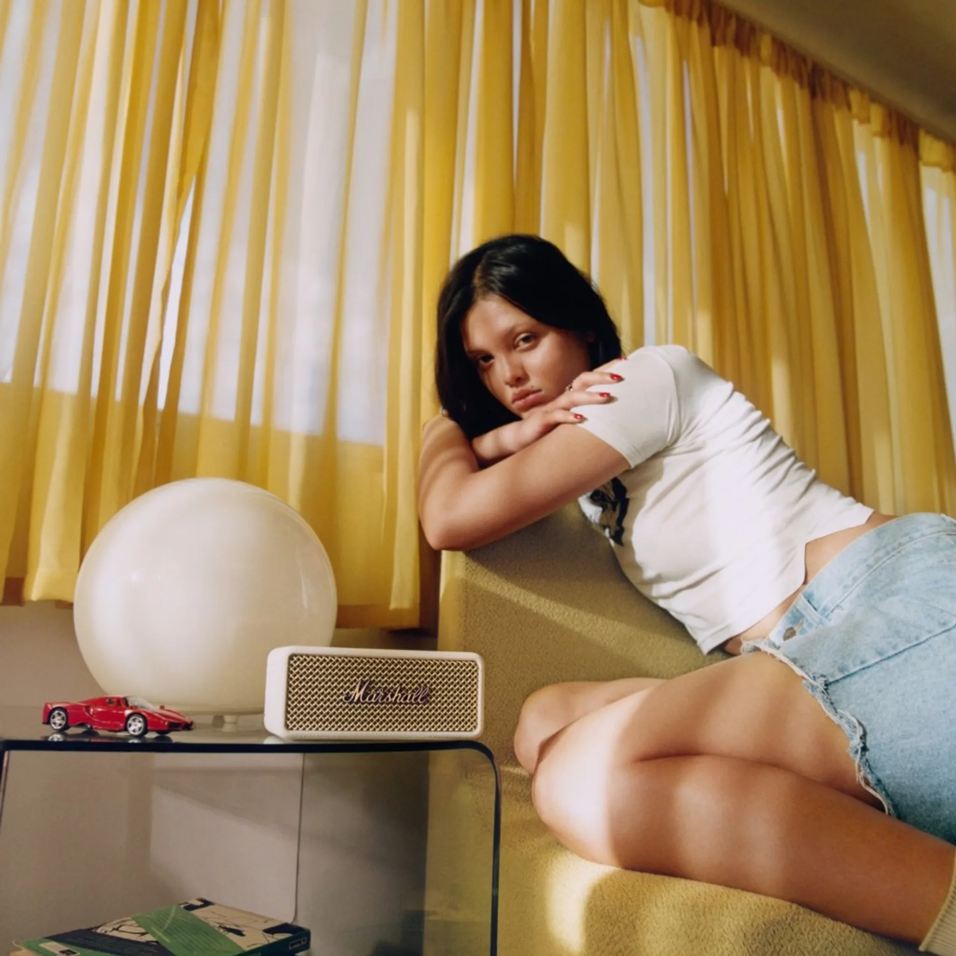 Lifestyle shot with woman by yellow curtains, glass table with Marshall Emberton III cream speaker, lamp, and red toy car.