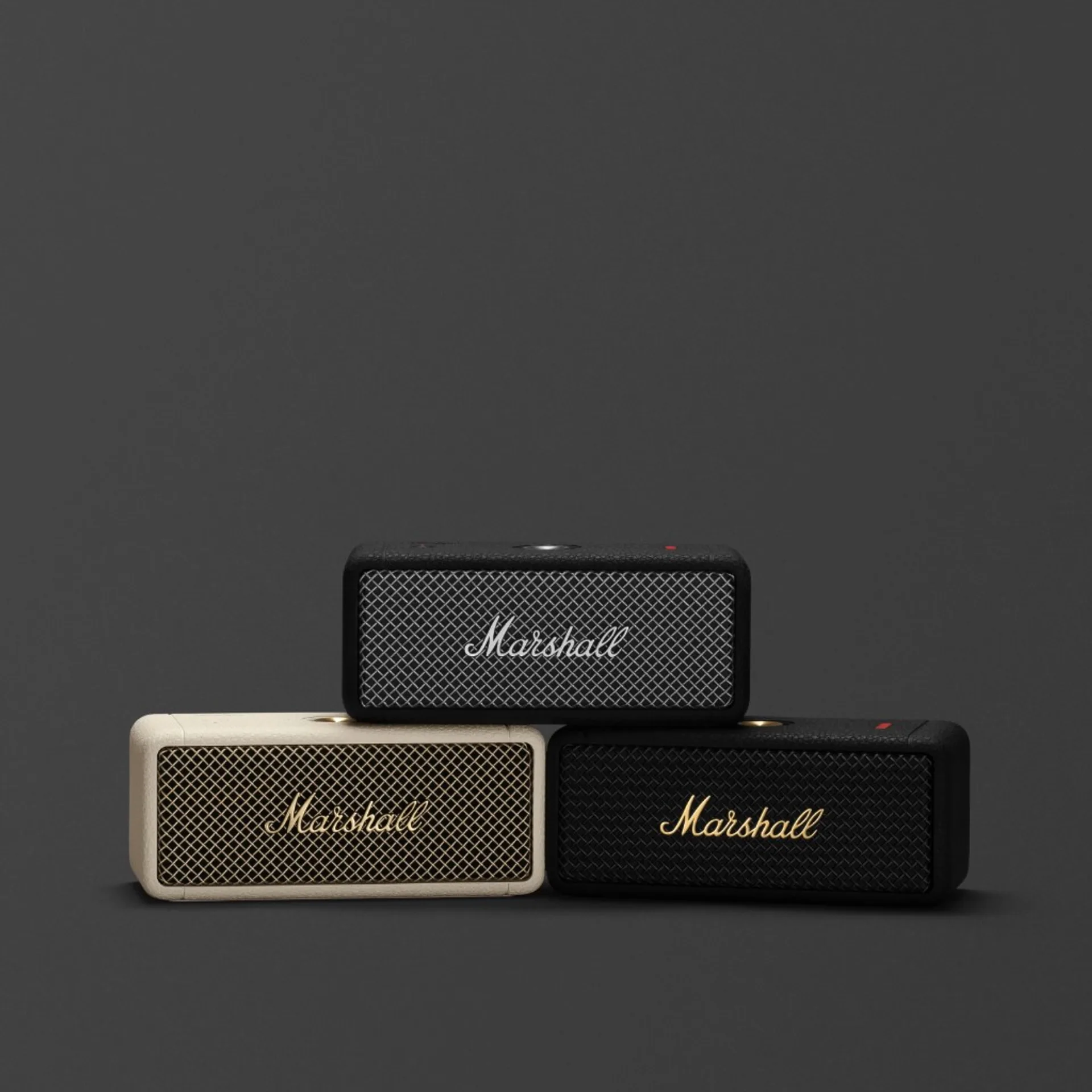 Three Marshall Emberton portable speakers stacked pyramid style in black, cream, and black-gold finishes on charcoal backgro…