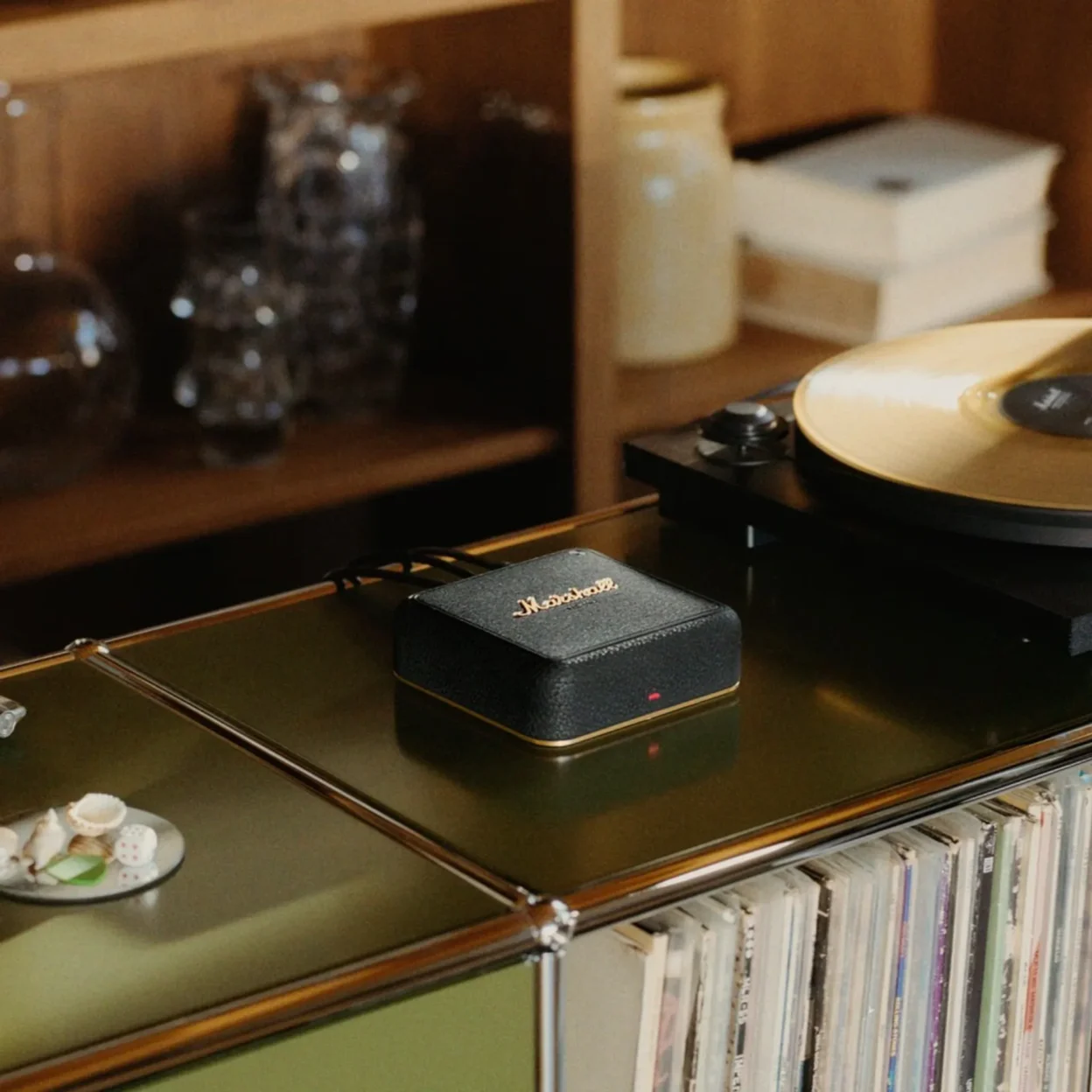 Small black Marshall device with gold logo on olive-green glass shelf beside turntable with gold vinyl, records and decor in…