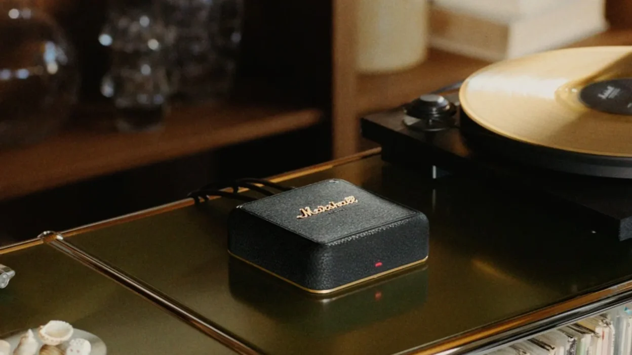 Small black Marshall device with gold logo on olive-green glass shelf beside turntable with gold vinyl, records and decor in…