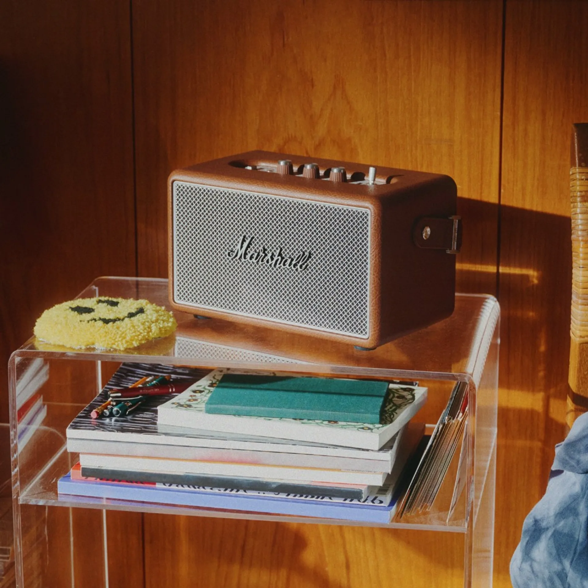 Brown Marshall Kilburn III on clear acrylic table with smiley rug, notebooks, and sunlit wood panel wall.