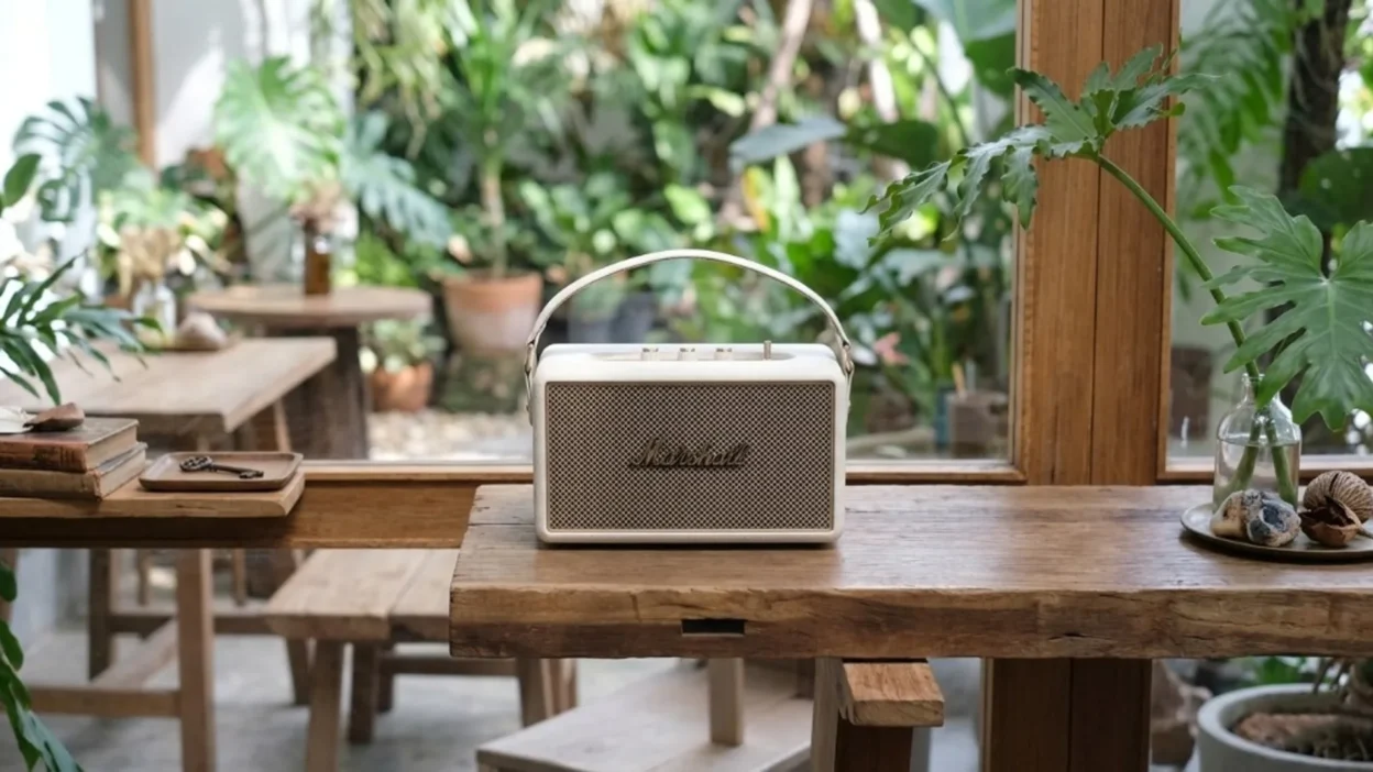 Marshall Kilburn III in cream finish with gold grille and carry strap on a rustic wood table, bright cafe interior with tropical plants.