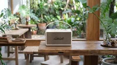 Marshall Kilburn III in cream finish with gold grille and carry strap on a rustic wood table, bright cafe interior with tropical plants.